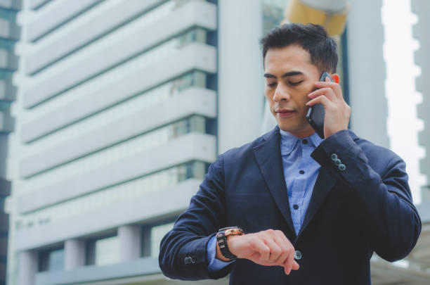 portrait attractive handsome businessman using smartphone talking and looking smart watch checking time to working against high building background.