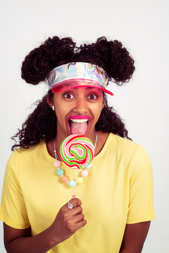 Young funny woman with pink lipstick licking the spiral lollipop, looking at a camera. The woman wearing a yellow t-shirt, sun visor. Head and shoulders portrait of elated beautiful African female model on white background. Purple film filter applied.