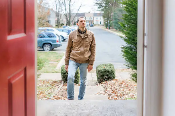 Young man standing on front porch frontyard steps in front of house home buyer looking to buy property real estate customer client in townhouse residential neighborhood, cars parked, door open
