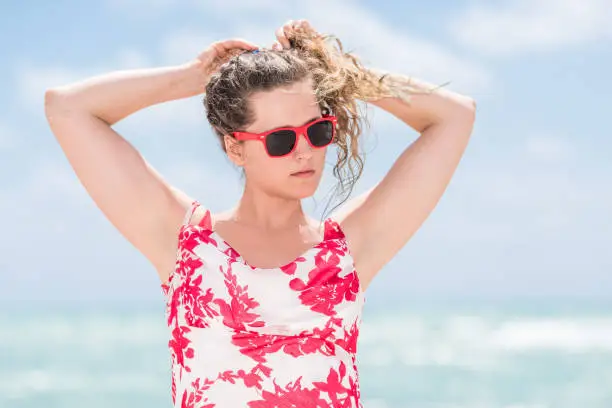 Young woman hipster millennial in dress on beach during sunny day with red sunglasses in Miami, Florida with ocean background, tying hair arms