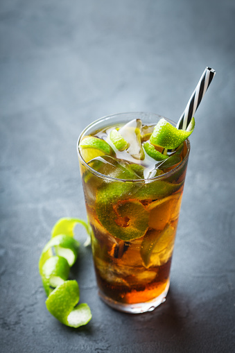 Food and drink, holidays party concept. Alcohol cocktail with rum and cola cuba libre beverage, longdrink in a glass with straw, ice and lime zest on a dark table