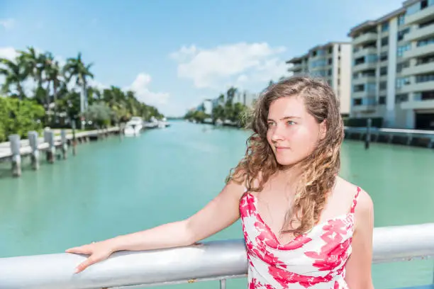Young woman hipster millennial in red dress during sunny day in Bal Harbour, Miami, Florida with green ocean Biscayne Bay background, leaning on bridge railing