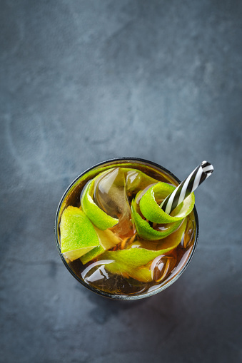 Food and drink, holidays party concept. Alcohol cocktail with rum and cola cuba libre beverage, longdrink in a glass with straw, ice and lime zest on a dark table. Copy space top view background