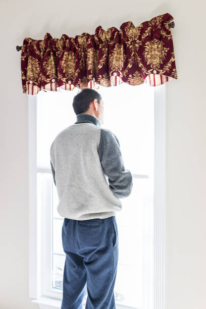 istock Young man standing in front of bright window in room adjusting old-fashioned retro blinds curtains 968533436