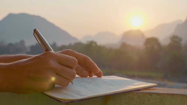 Young woman writing morning pages in diary outdoor, close-up Young woman writing travel notes in diary during sunrise with beautiful sun light and mountain landscape on the background, close-up. diary stock pictures, royalty-free photos & images