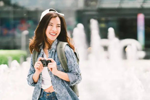 Photo of Young beautiful Asian backpack traveler woman using digital compact camera and smile, looking at copy space. Journey trip lifestyle, world travel explorer or Asia summer tourism concept