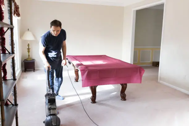Young man house husband stay at home dad vacuuming using vacuum on carpet floor inside interior of house living room billiards table, domestic life