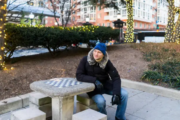 Empty chess table in park in Washington DC city capital with young man sitting smiling in evening winter holiday lights, cold day, coat