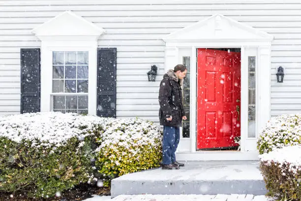 Photo of Young man outside front yard red door of house with snow during blizzard white storm, snowflakes falling letting calico cat outside outdoors to porch