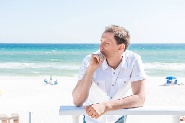 istock Young handsome attractive man serious on beach during sunny day in Seaside, Florida panhandle town village with ocean, hand on chin, leaning on railing, white shirt 968626374