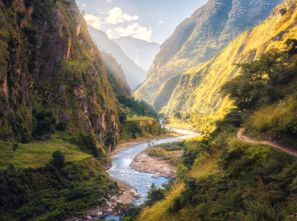 paysages colorés avec hautes montagnes de l’himalaya, belle rivière courbante, verte forêt, bleu ciel avec nuages et le soleil jaune au coucher du soleil en été au népal. vallée de montagne. voyage dans l’himalaya - fleuve et rivière photos et images de collection