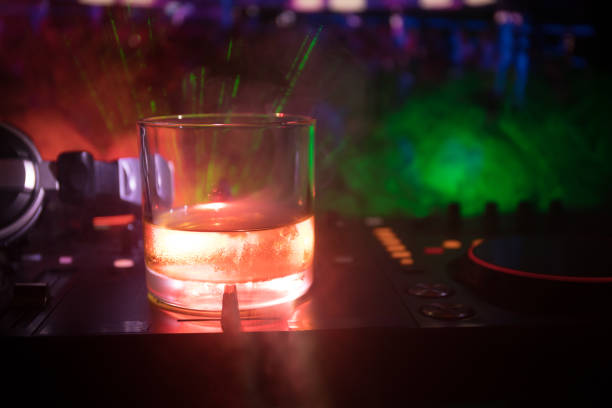 istock Glass with whisky with ice cube inside on dj controller at nightclub. Dj Console with club drink at music party in nightclub with disco lights. 968636362