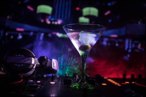 istock Glass with martini with olive inside on dj controller in night club. Dj Console with club drink at music party in nightclub with disco lights. Close up view 968636378