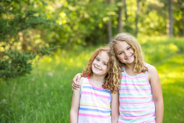 Close-up of two young girls (sisters) standing side by side on a summer day. They are on a hike through the woods and have stopped to take a picture. Both girls are smiling at the camera, and the older sister has her arm around the younger sister. The younger sister is on the left with curly red hair. The older sister is on the right with curly blonde hair.