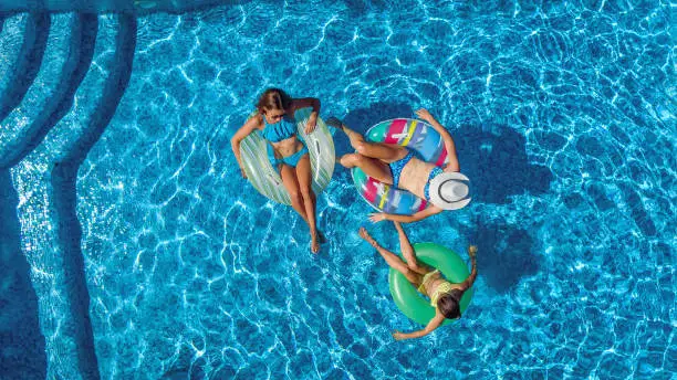 Photo of Aerial top view of family in swimming pool from above, happy mother and kids swim on inflatable ring donuts and have fun in water on family vacation