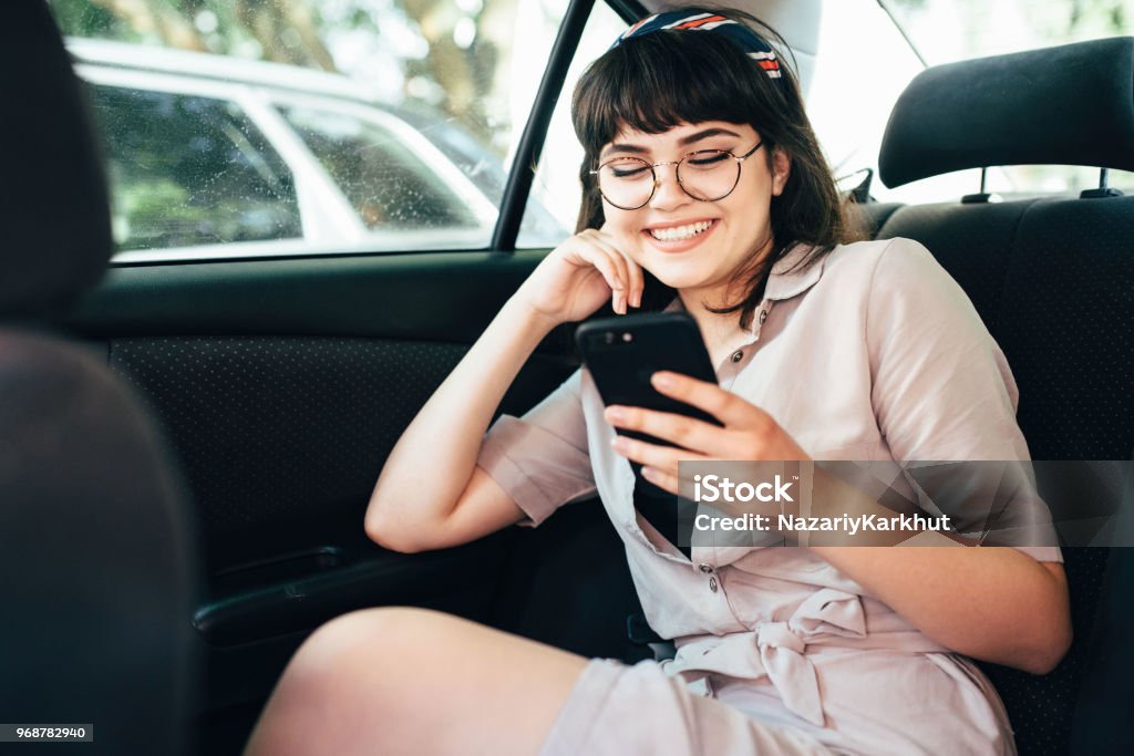 Beautiful young pretty girl is using a smart phone and smiling while sitting on back seat in the car Beautiful young pretty girl is using a smart phone and smiling while sitting on back seat in the car. Car Stock Photo