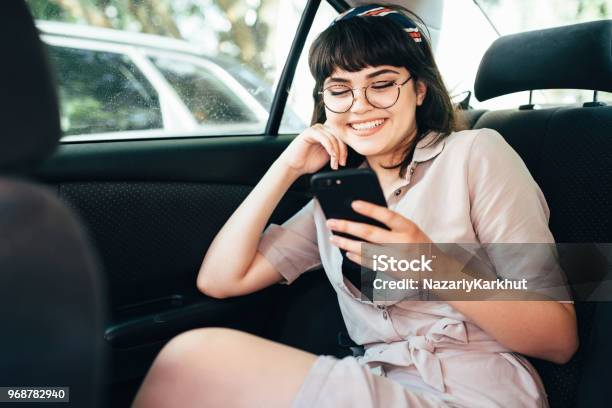 Beautiful Young Pretty Girl Is Using A Smart Phone And Smiling While Sitting On Back Seat In The Car Stock Photo - Download Image Now