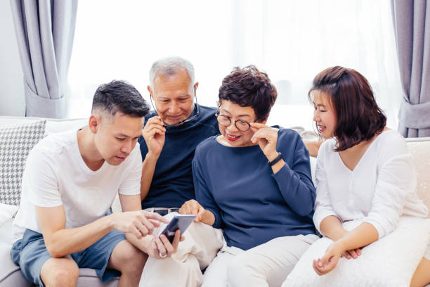 asian family with adult children and senior parents using a mobile phone and relaxing on a sofa at home together - father digital tablet asian ethnicity daughter imagens e fotografias de stock