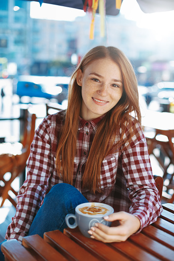 Coffee concept. Natural reddish girl with freckles, dressed in plaid shirt, smiling while holding a big cup of coffee latte. Portrait of attractive young woman drinking coffee outdoors.