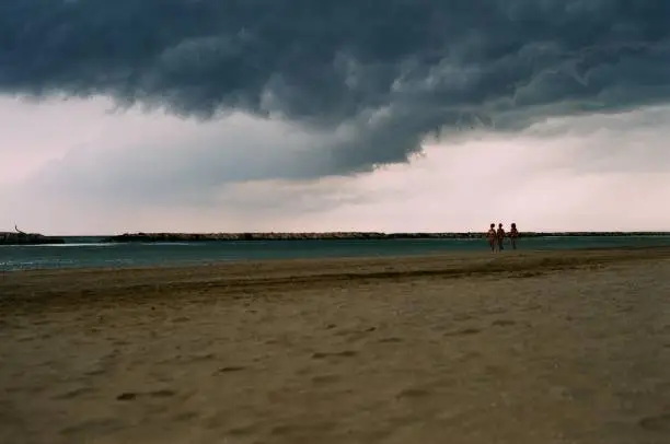 Dramatic sky with dark clouds, the light under and small figures walking along the shore. Analog film photo