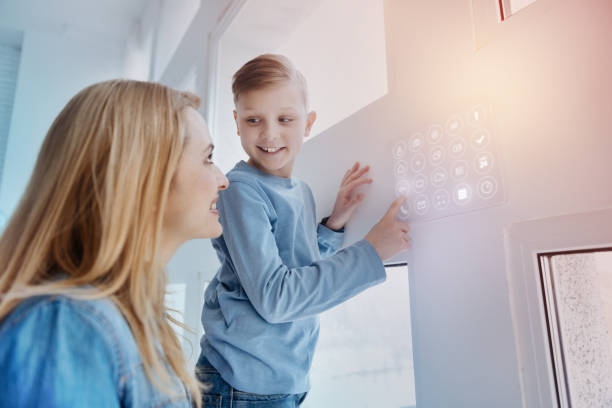 Smiling boy looking at his mother while touching a device on the wall Curious child. Cheerful kind boy touching a modern device on the wall of his smart house and looking attentively at his mother smart home family stock pictures, royalty-free photos & images