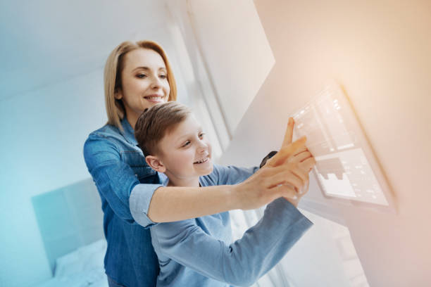 Smiling boy pressing his finger to the screen and his mother helping him Funny device. Cheerful emotional boy feeling excited while his kind mother helping his to use a modern device on the wall smart home family stock pictures, royalty-free photos & images