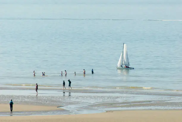 people on the beach watching a sailing boat a coasting near the sand at springtime and some others enyoing the first warmth in the water