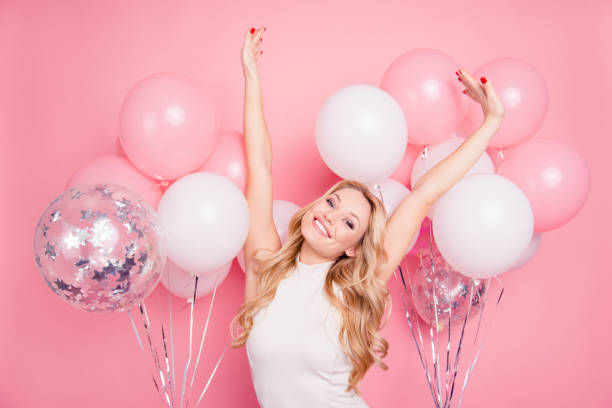 Portrait of funky glad girlfriend with beaming smile raised hands, making stretching having many white and pink air balloons on background