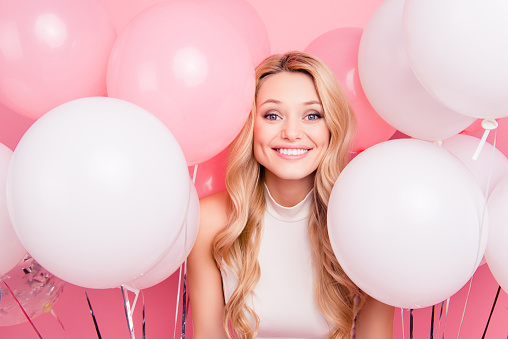 Portrait of cheerful childish girl with many white air balloons around looking at camera isolated on pink background