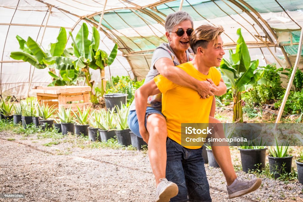 grandmother and nephew teenager play together doing horse transportation in a plants shop outdoor. sunny day and happiness having fun concept for different generations and ages. smiles and laugh Grandmother Stock Photo