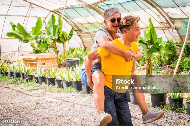 Grandmother And Nephew Teenager Play Together Doing Horse Transportation In A Plants Shop Outdoor Sunny Day And Happiness Having Fun Concept For Different Generations And Ages Smiles And Laugh Stock Photo - Download Image Now