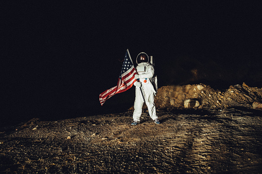 One man, astronaut exploring the land on the other planet alone at night, holding a USA national flag.