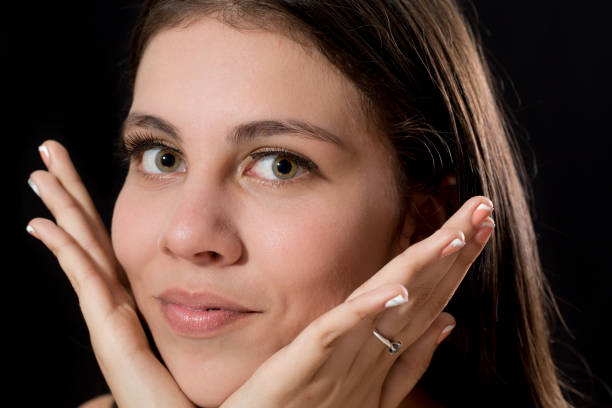 Beautiful seventeen years old latin lady in a studio portrait, She is looking at the camera, She is serious, She has long smooth hair and green eyes. She is wearing a V-neck cream color blouse. The background is black. This is an extreme close-up headshot. She has her hands under her chin holding her face.