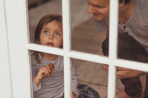 Little girl sitting beside a window and looking outside holding a piece of cake. Man sitting beside her little daughter near a window.