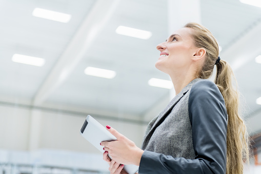 Horizontal color image of pretty businesswoman in warehouse holding touchpad, looking up to the ceiling and smiling.