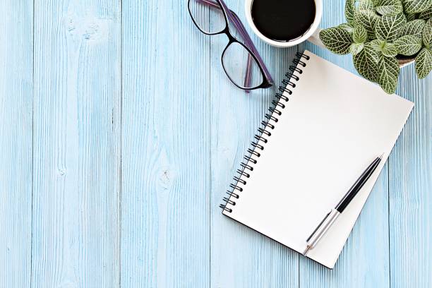 istock Open notebook with blank pages, coffee cup and eyeglasses on wooden desk table, top view or flat lay with copy space ready for adding or mock up 968977150