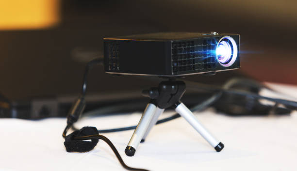 istock Black projector with tripod installed on white table, in hall or seminar meeting room. Real projector at business conference or presentation in the office room. Close up of projector for cinema and video projections. Business and education concept. 969039554