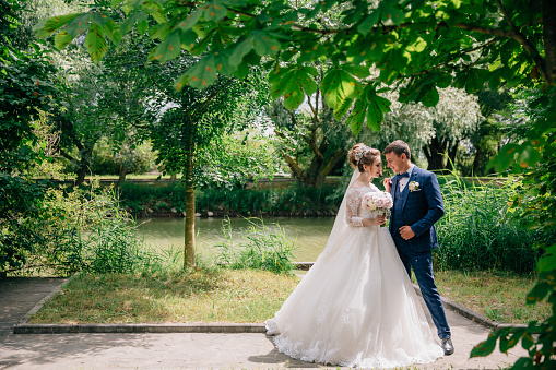Newlyweds are walking among the green trees in the park. The bride in a snow-white lush dress and with a gently pink bouquet of pions hugs her fianc in a stylish suit