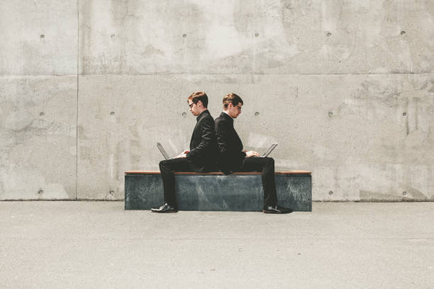 twin brothers working with laptop concept twin brothers with black suits and glasses sitting back on back on a bench while working on their laptop. two men same clothes stock pictures, royalty-free photos & images
