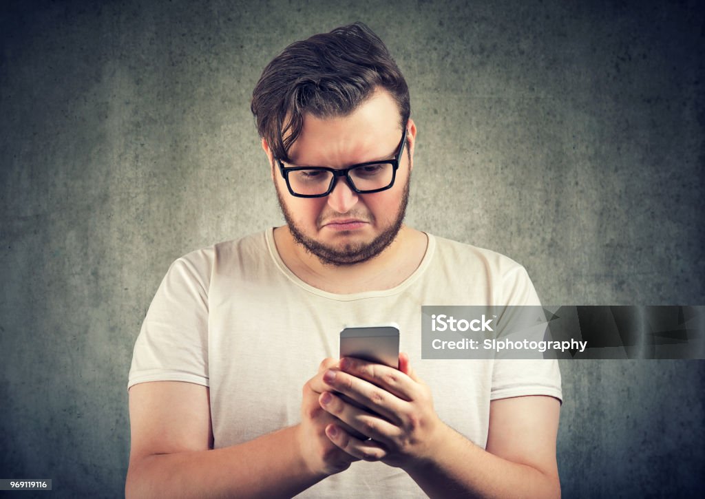 Upset man watching smartphone Casual sad young man received offensive message on smartphone looking upset and gloomy Disgust Stock Photo