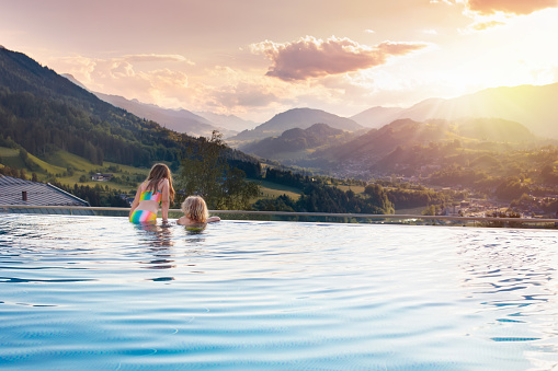 Kids play in outdoor infinity swimming pool of luxury spa alpine resort at sunset in Alps mountains. Spring or summer vacation for family with children. Boy and girl in hot tub with mountain view.