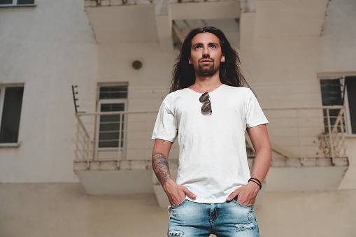 Portrait confident man standing in powerful pose, looking straight holding hands in pockets on building background. Young tattooed casual man with long hair, jeans and white t-shirt.