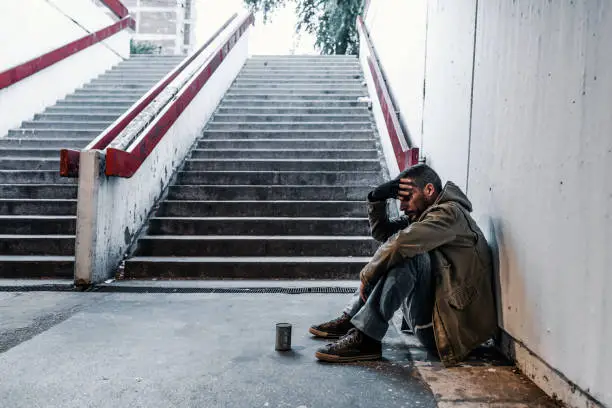 Photo of homeless man sitting in the train station, Subway station, tunnel or underground and asking for help with donation can near him. Side view of Desperate mature man who lost job abandoned and lost in depression sitting on ground street wall suffering emotional pain.