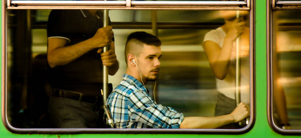 Young man with earphones in plaid shirt sitting while riding in a window seat of a tram  from outside stock photo