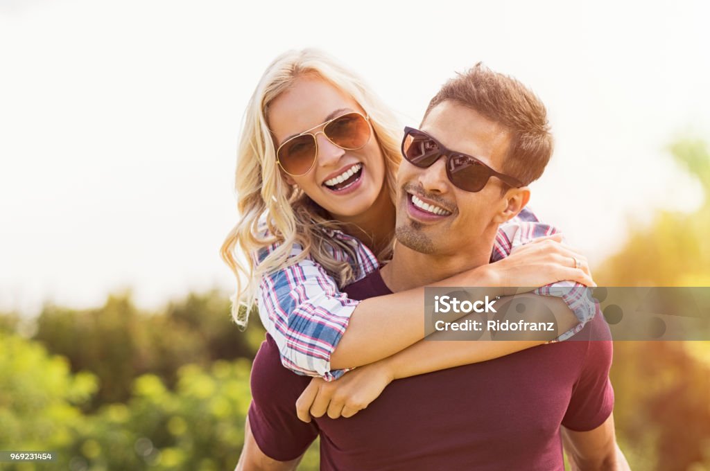 Man giving piggyback ride to woman Closeup of young man carrying beautiful woman on his back. Smiling girl piggyback on guy while looking at camera. Boyfriend giving girlfriend piggyback ride and smiling outdoor. Couple - Relationship Stock Photo