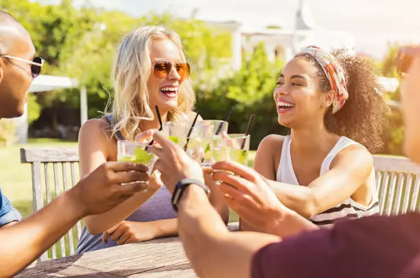 Two happy young couples toasting with glasses of cocktails. Young beautiful women and men drinking lemonade and smiling. Cheerful guys and multiethnic girls friends raising a mojito toast at summer party."r