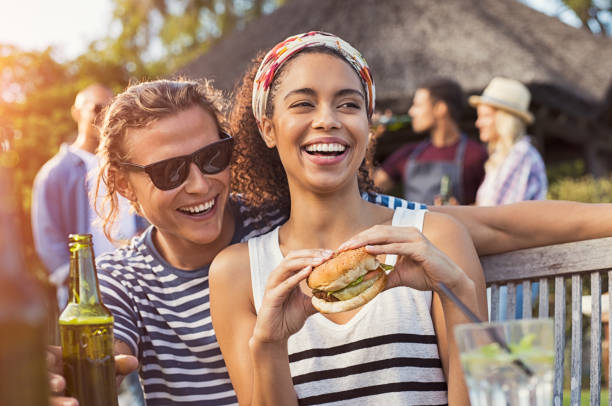 Couple enjoying food and drinks at party Cheerful multiethnic couple enjoying hamburger and beer at barbecue party. Happy young black woman eating burger while is sitting on boyfriend legs. Joyful guy and girl having fun at bbq party. dinner party photos stock pictures, royalty-free photos & images
