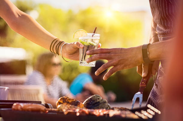 Woman sharing drink at bbq Closeup of young woman hand passing a drink to a man while preparing barbeque. Girl sharing glass with fresh lime juice during outdoor bbq. Hand of man taking mojito while preparing beef on bbq. south african braai stock pictures, royalty-free photos & images