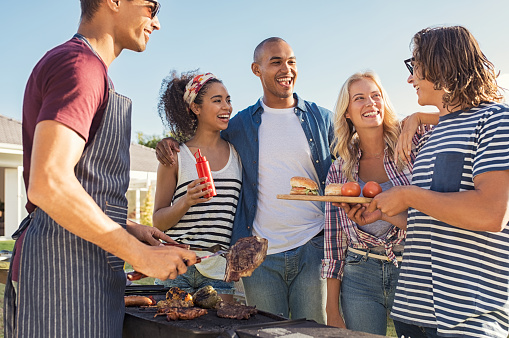 Young multiethnic friends having fun grilling meat enjoying bbq party. Group of happy guys and girls cooking and eating at barbecue dinner outdoor. Men and women standing around grill, chatting, drinking and eating.