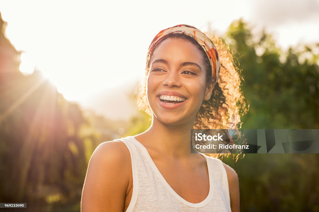 Young african woman smiling at sunset Portrait of beautiful african american woman smiling and looking away at park during sunset. Outdoor portrait of a smiling black girl. Happy cheerful girl laughing at park with colored hair band. Women Stock Photo